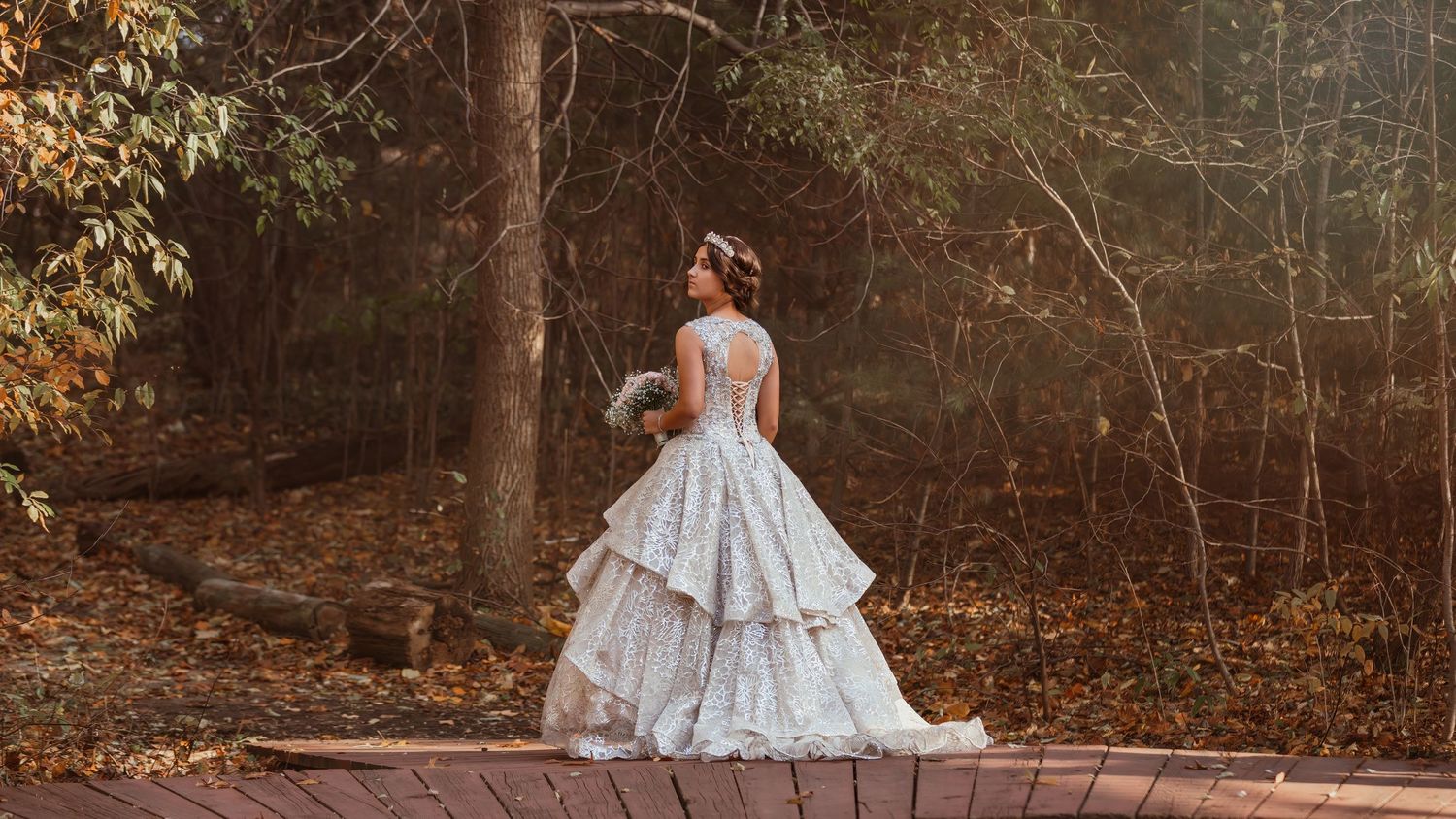 Bride in tiered wedding gown walking along pathway surrounded by autumn foliage.