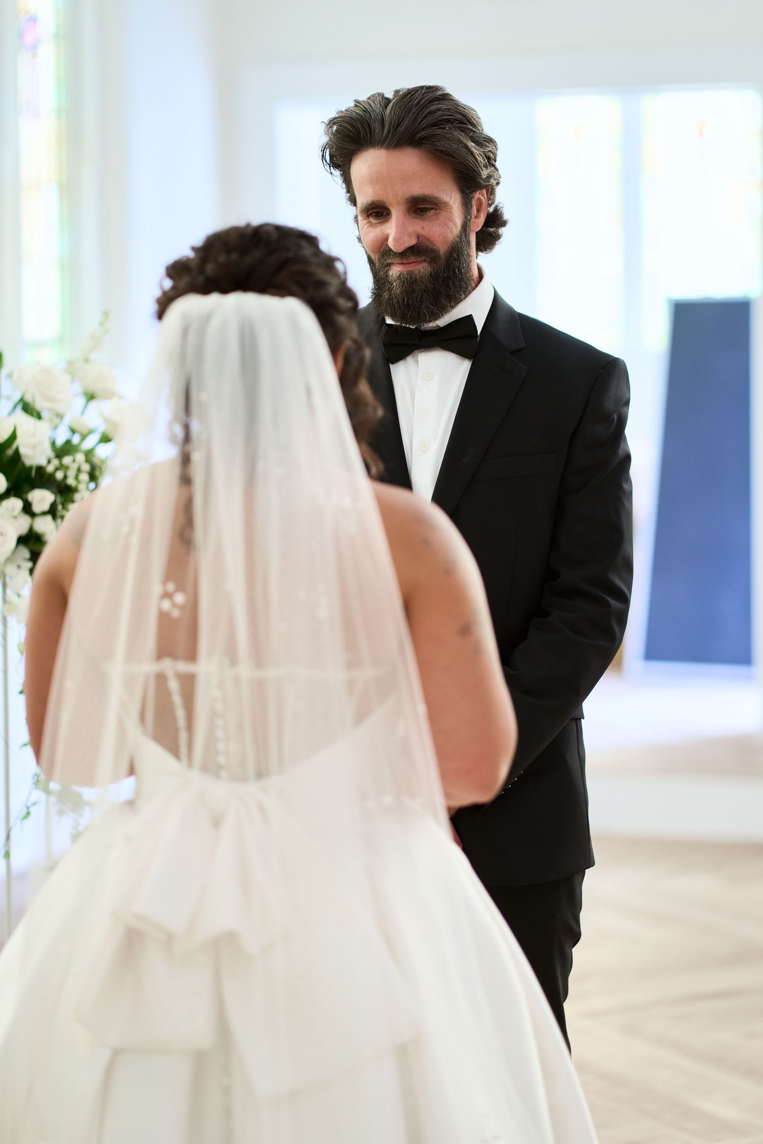 A wedding ceremony moment in a bright sunlit room with a bride in white and groom in black tuxedo.