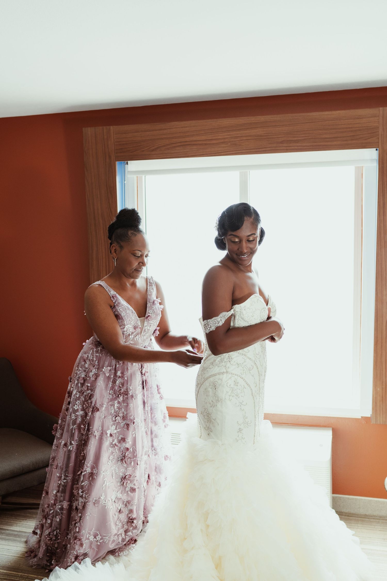 Two people in formal dresses preparing for a special occasion near a window.