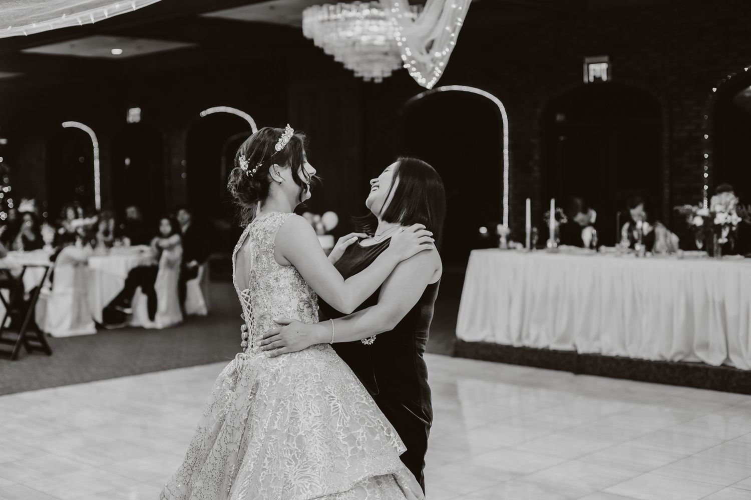 A black and white capture of two people dancing at a wedding reception with white-draped tables in background.