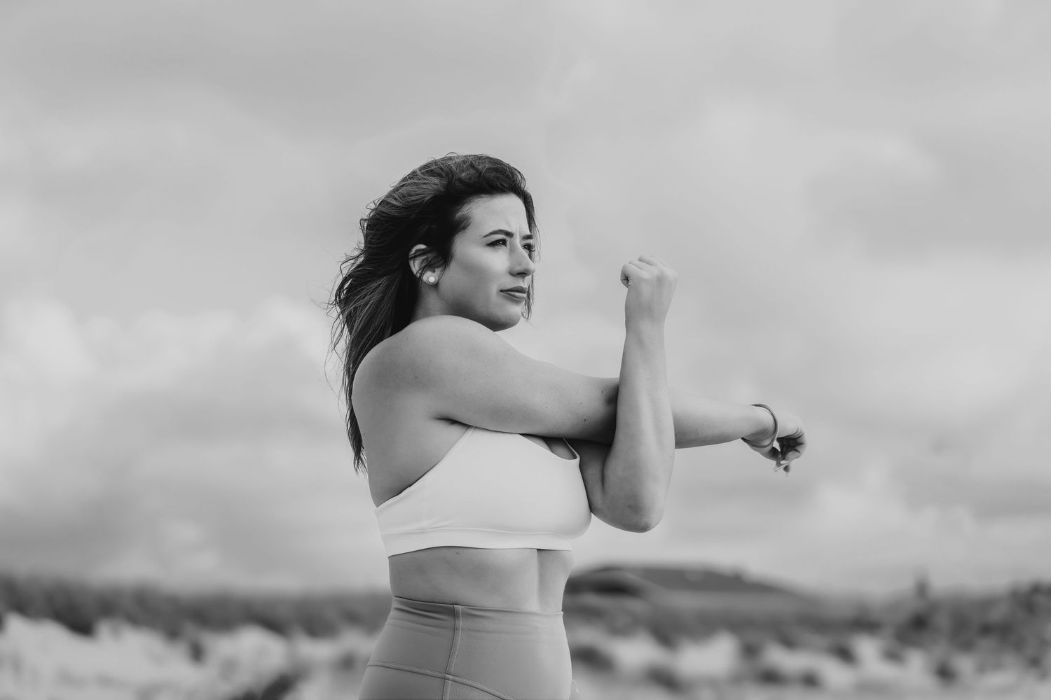 A woman in athletic wear stretches her arms during an outdoor workout against a cloudy sky.