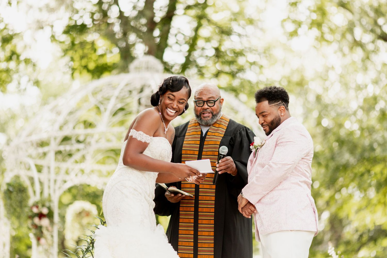 Wedding participants share laughter during their outdoor ceremony beneath green trees.