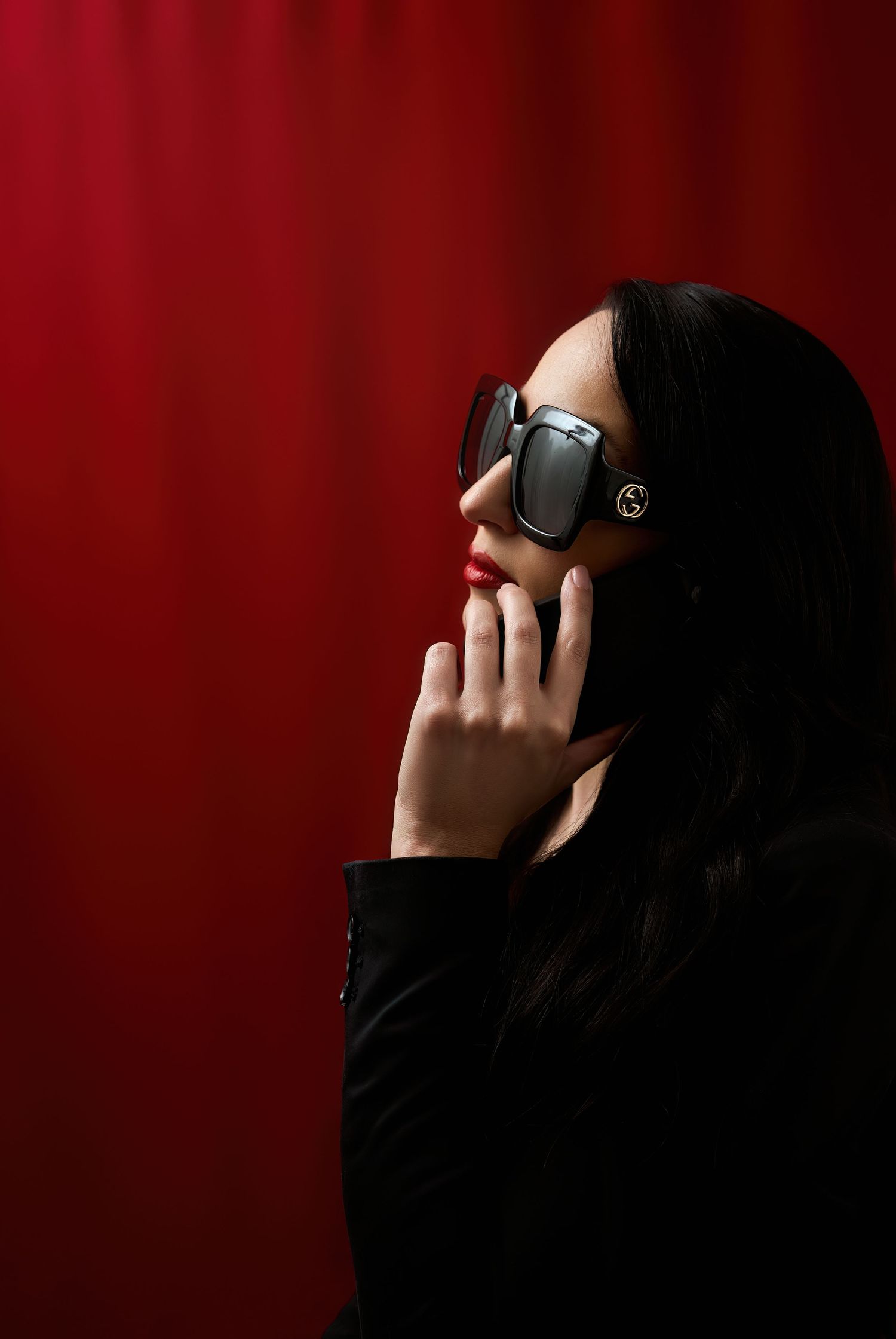 Stylish person in sunglasses and black outfit poses thoughtfully against a deep red background.