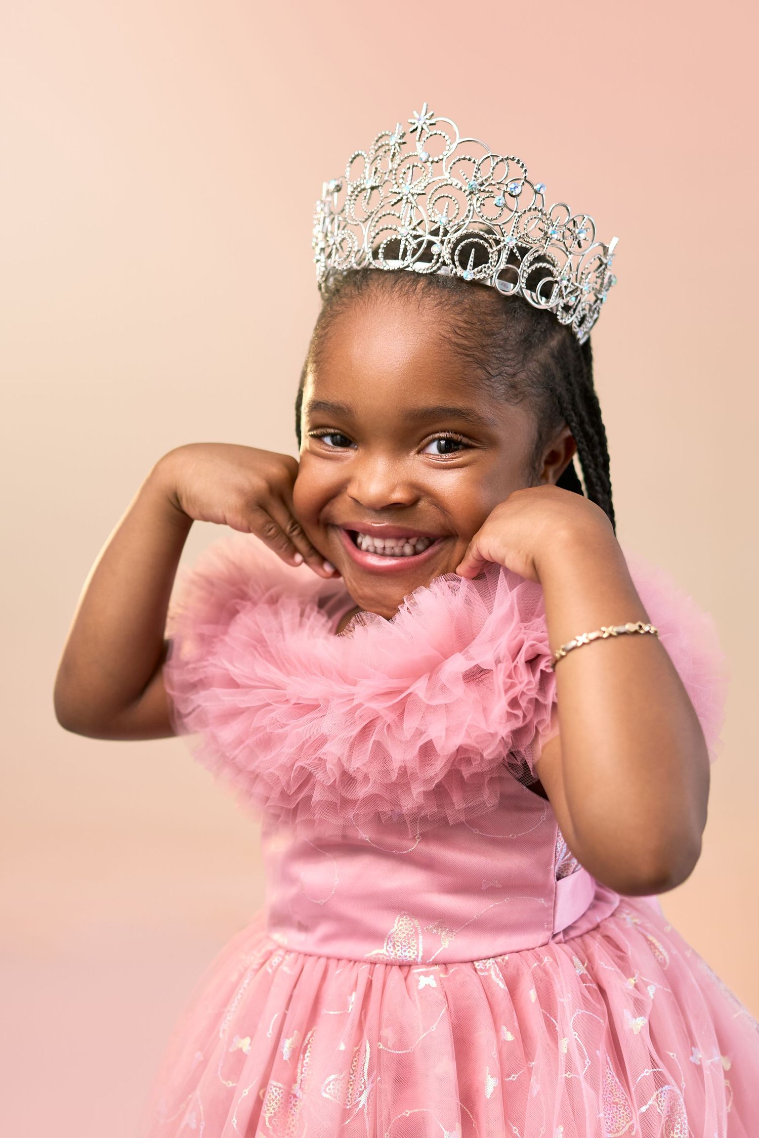 A young princess in a sparkly tiara and pink tulle dress poses against a pink background.