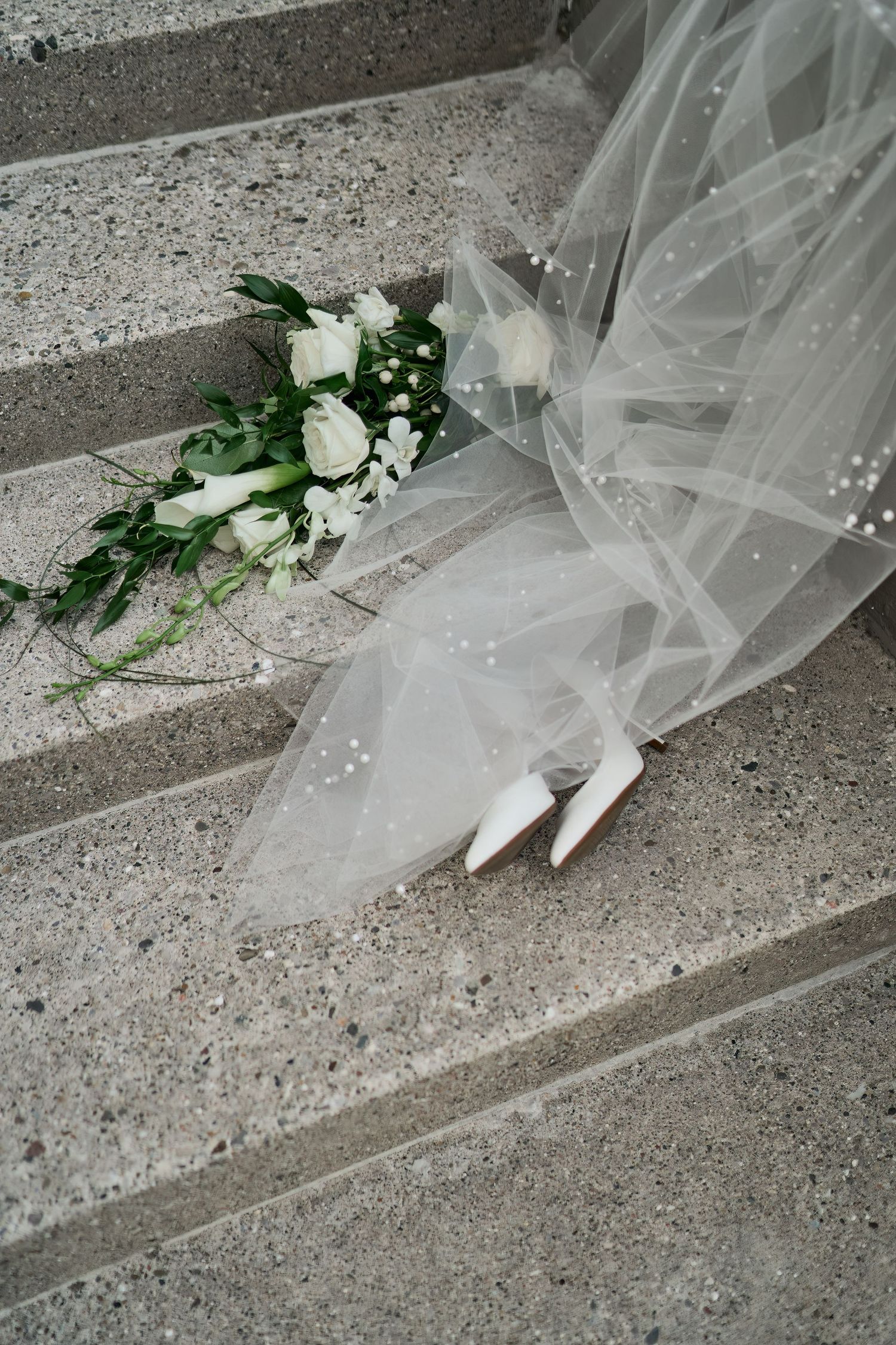 A white bridal bouquet and veil rest on concrete stairs in an elegant wedding detail shot.