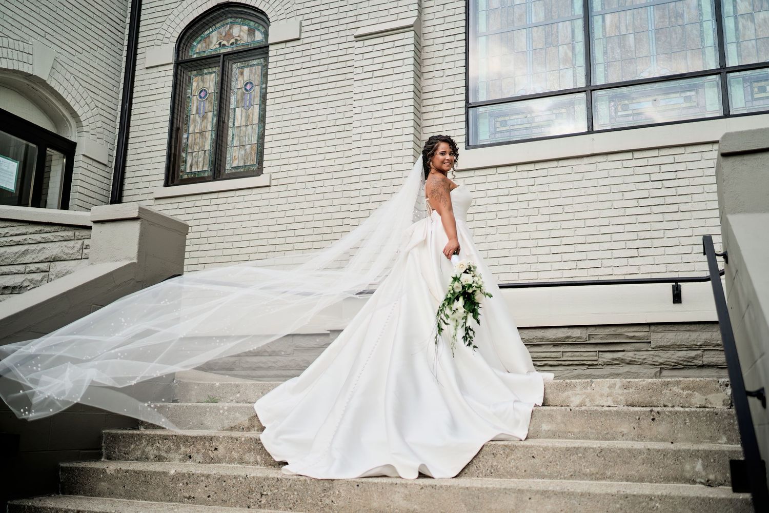 A bride in a flowing white wedding dress stands on stone steps holding a bouquet, with her long veil trailing behind her.