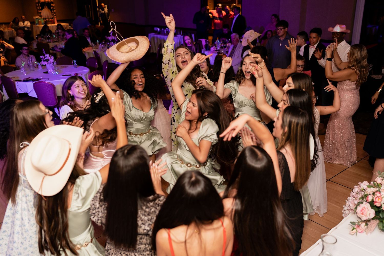 Wedding guests dance enthusiastically on a crowded dance floor under purple mood lighting at a reception celebration.