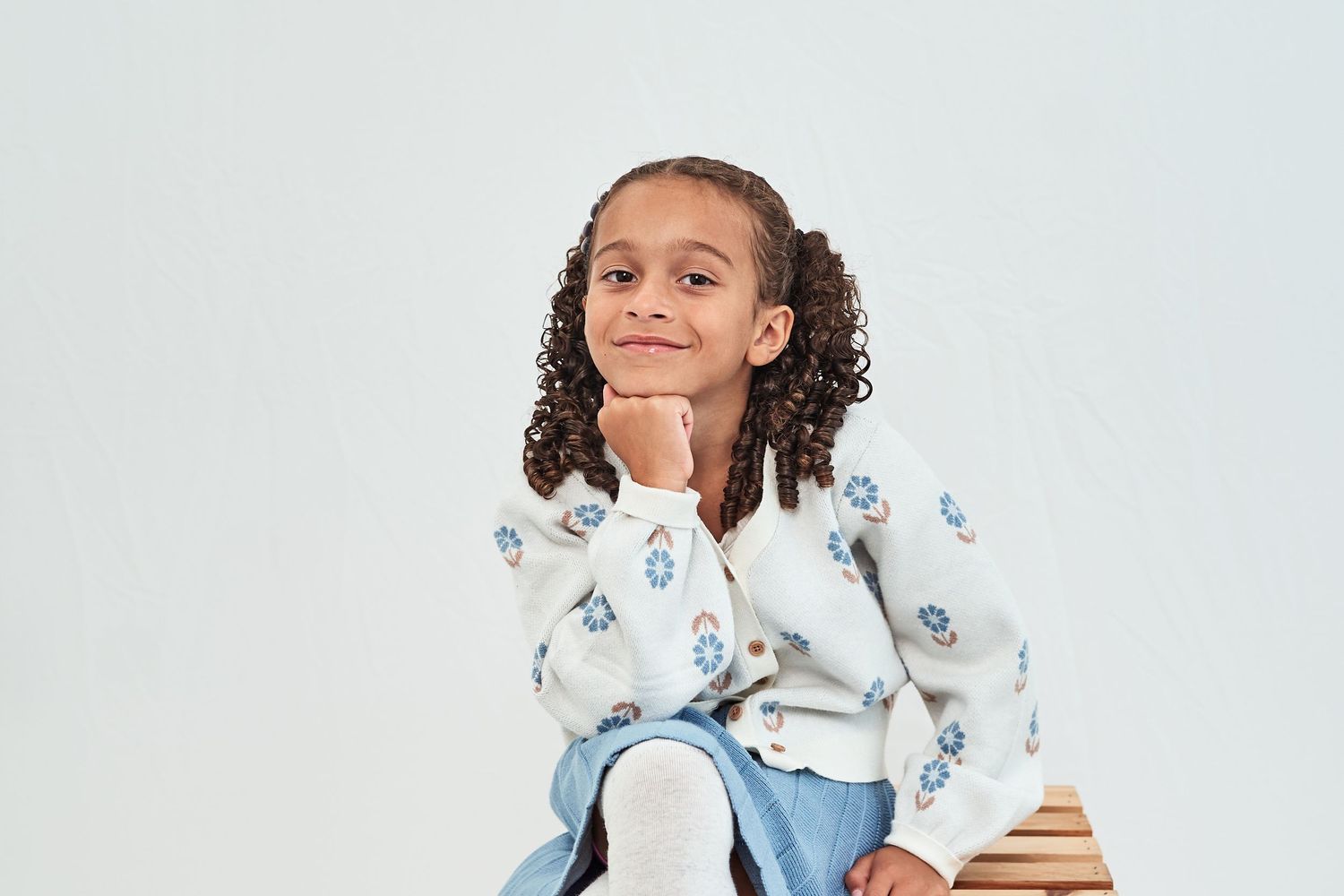 A young person with curly hair wearing a white and blue patterned top sits with a thoughtful expression.