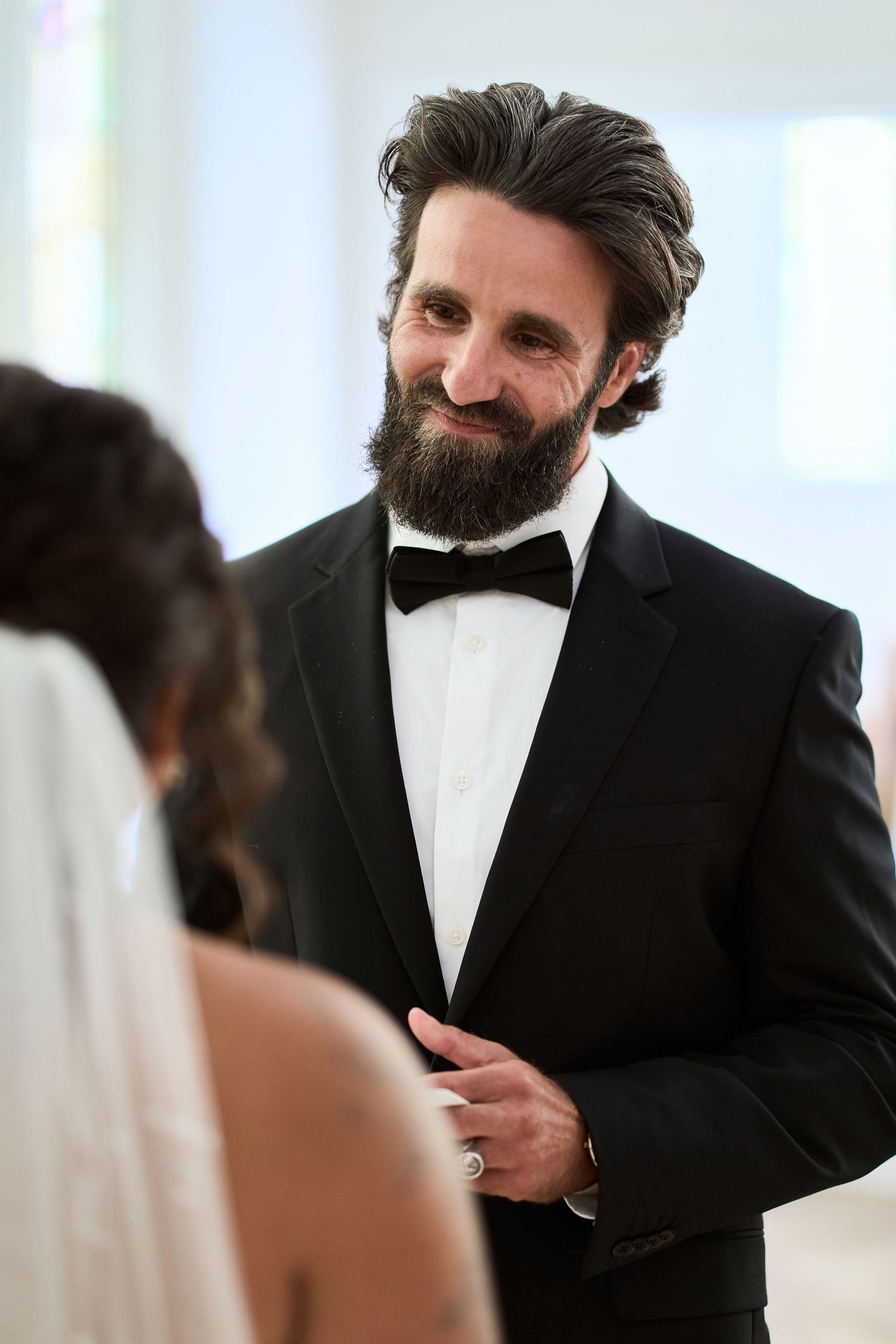 A person in a formal black tuxedo and white bowtie smiles during what appears to be a wedding ceremony.
