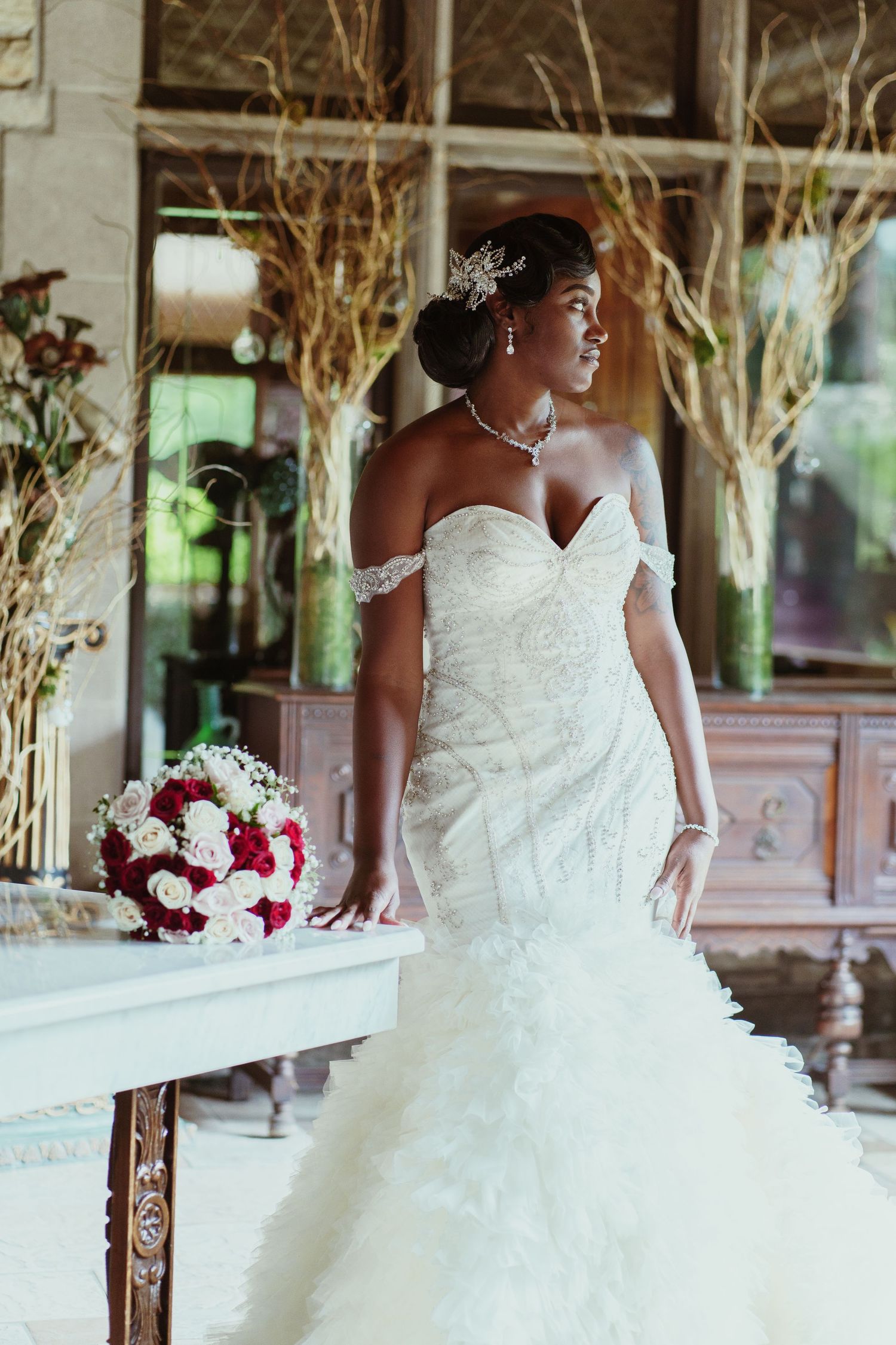 Elegant bride in off-shoulder mermaid gown poses with red and white bouquet in stone interior.