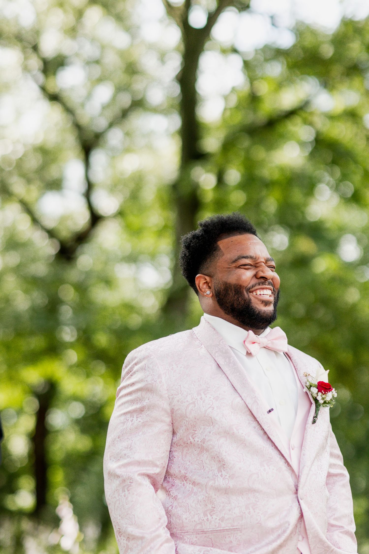 A person in a light pink tuxedo with a red boutonniere smiles outdoors against a blurred green background.
