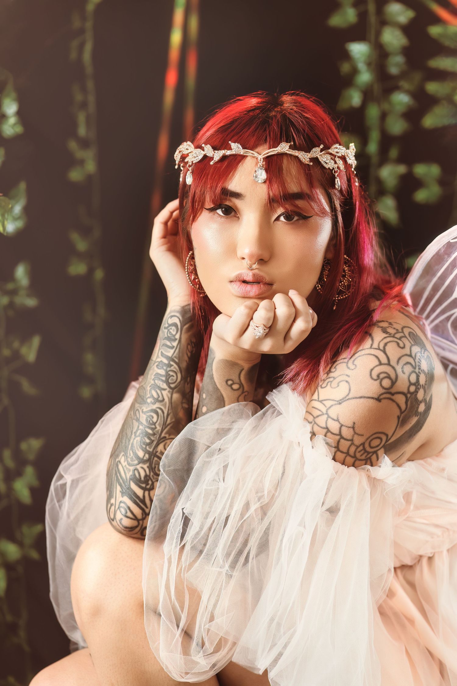 A person with red hair and tattoos poses in a dreamy ethereal white dress against a dark foliage background.