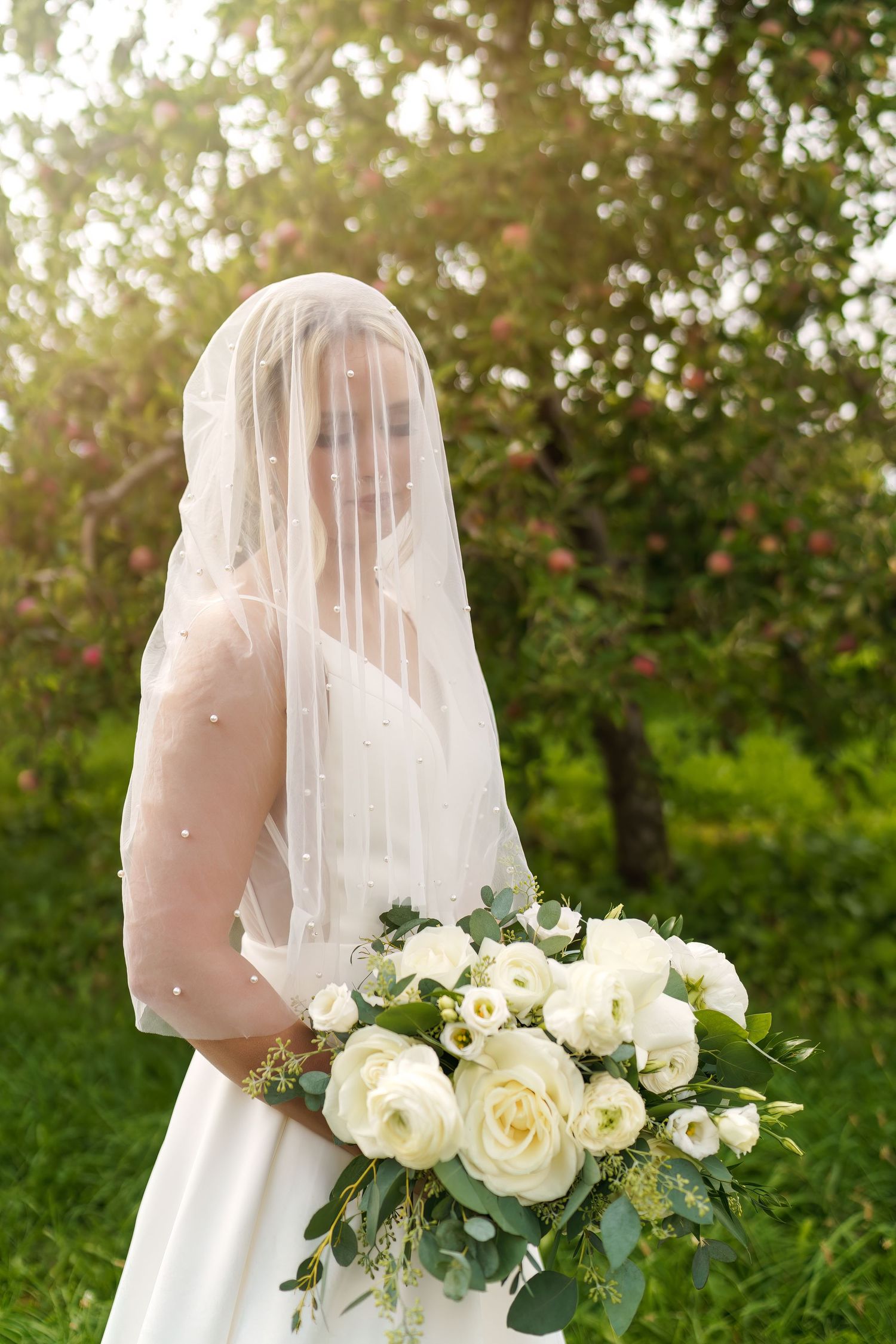 A bride in a white dress and veil holds a beautiful bouquet of cream roses and greenery in an outdoor garden setting.