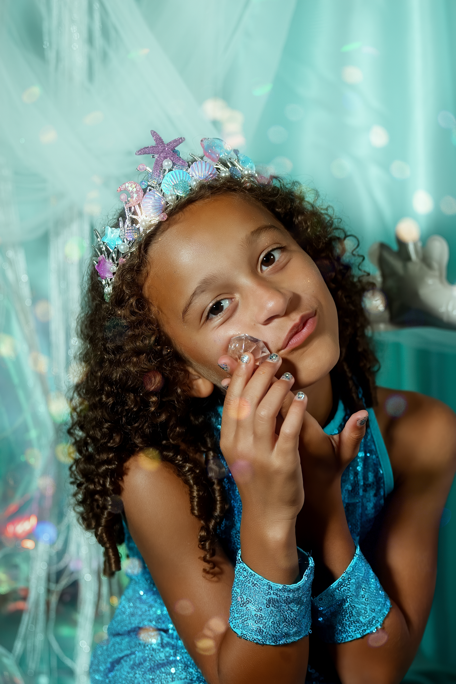 Young girl in blue sequin dress and mermaid crown poses dreamily with crystal prop in teal decorated setting.