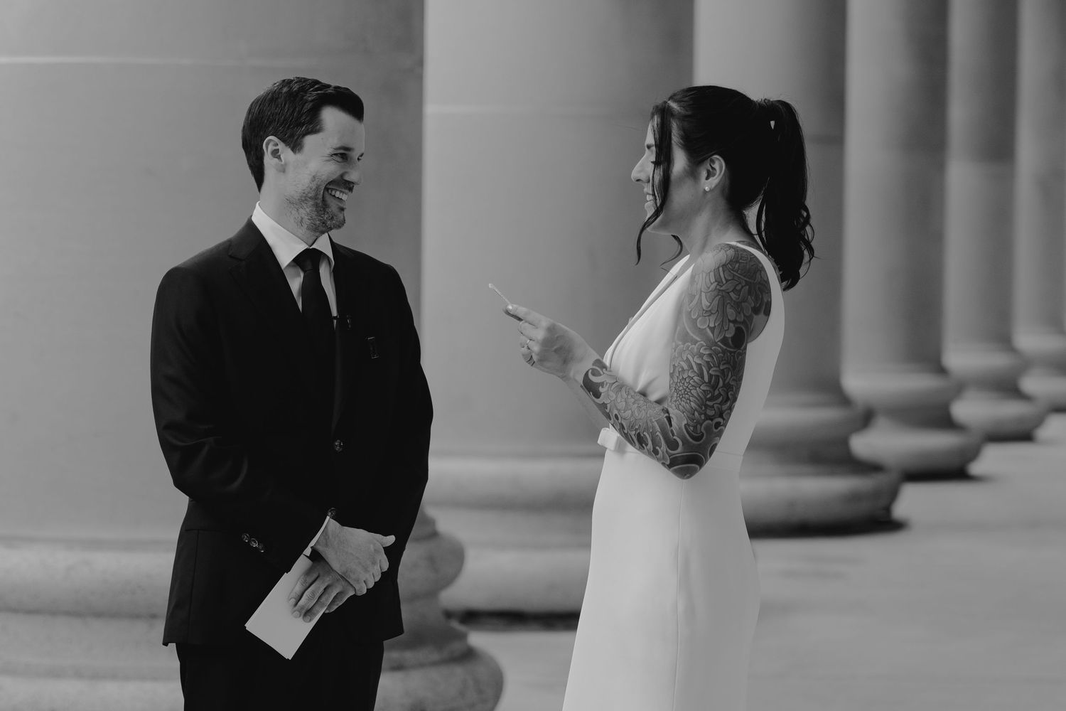 A bride and groom share a candid moment during their wedding ceremony in front of classical columns in black and white.