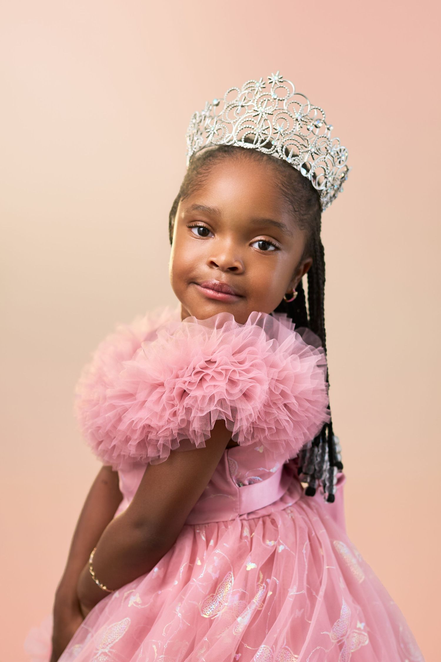 Young royal wearing an ornate silver tiara and pink tulle dress against a blush pink background.