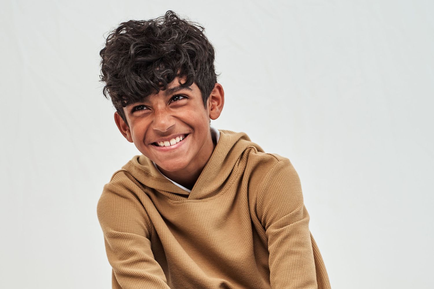 A young student wearing a tan hoodie smiles warmly against a light gray background.
