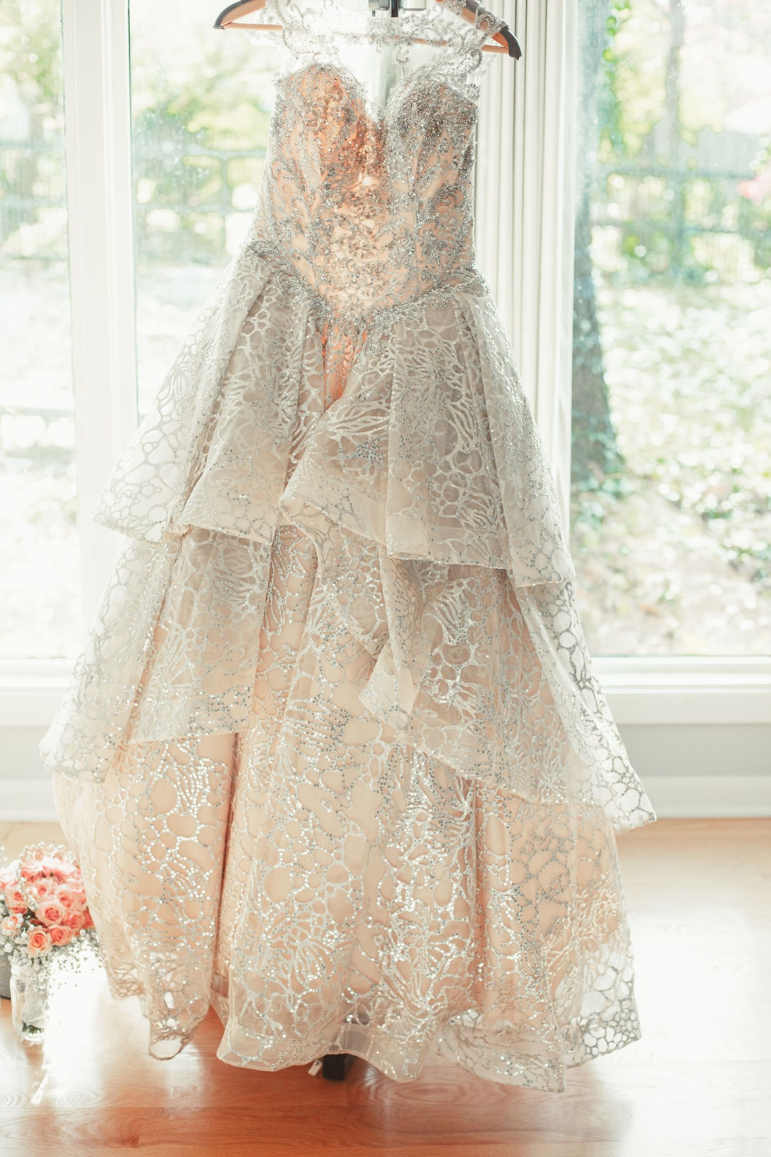 A luxurious beige lace wedding gown with tiered layers hangs elegantly by a window.