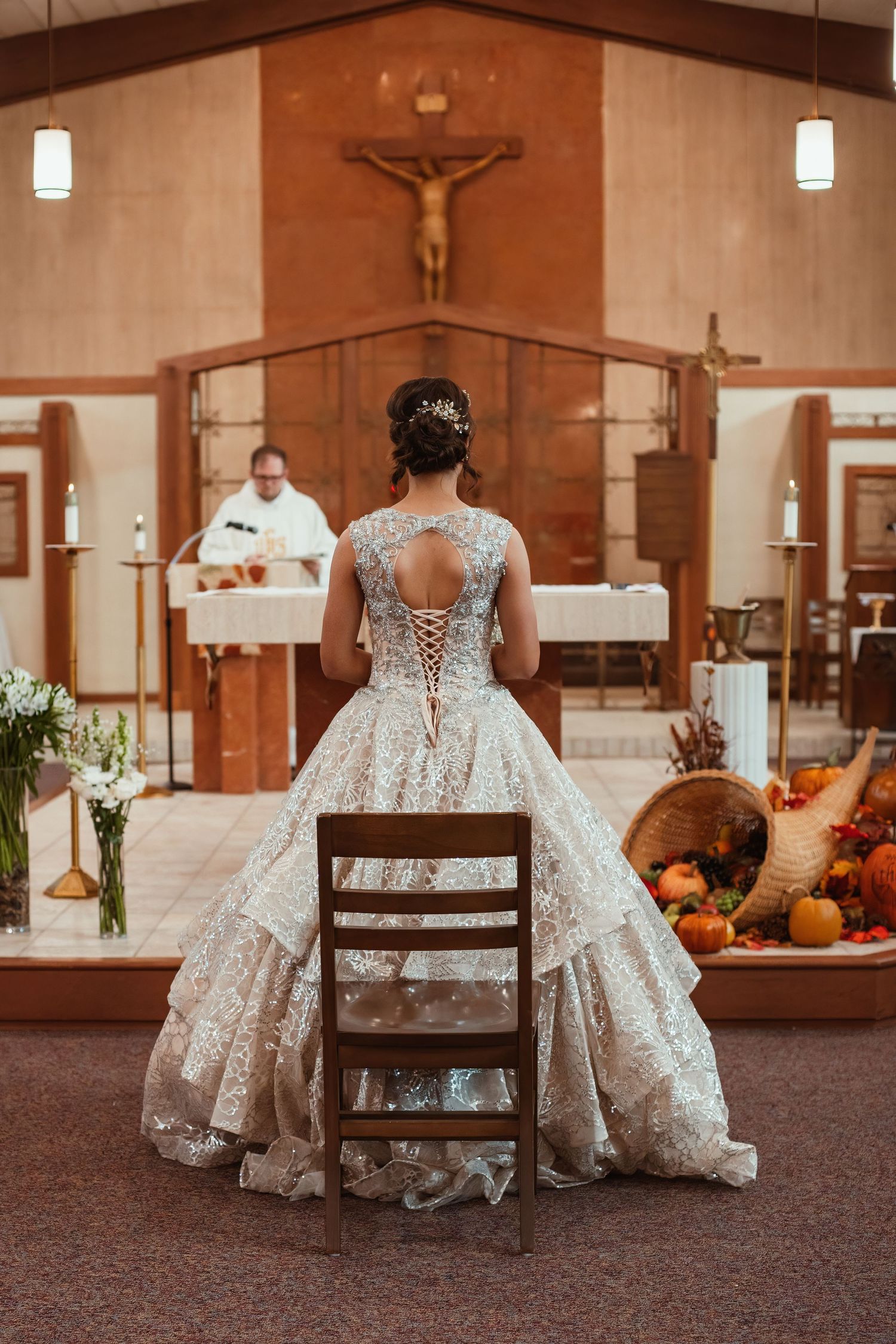 A layered lace wedding dress photographed from behind inside a church with a crucifix on display.