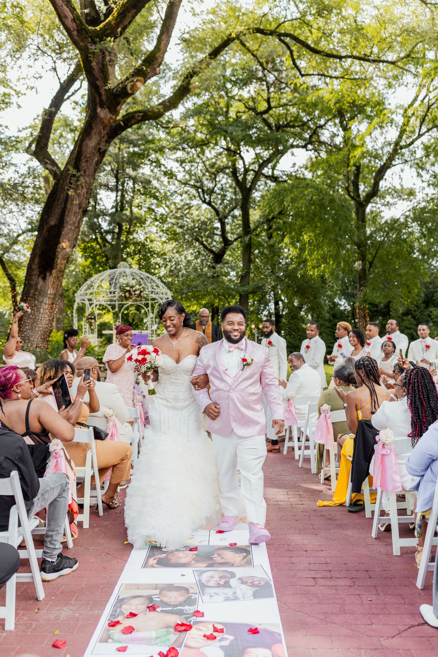 Newlyweds walk down decorated aisle while guests celebrate at garden ceremony.