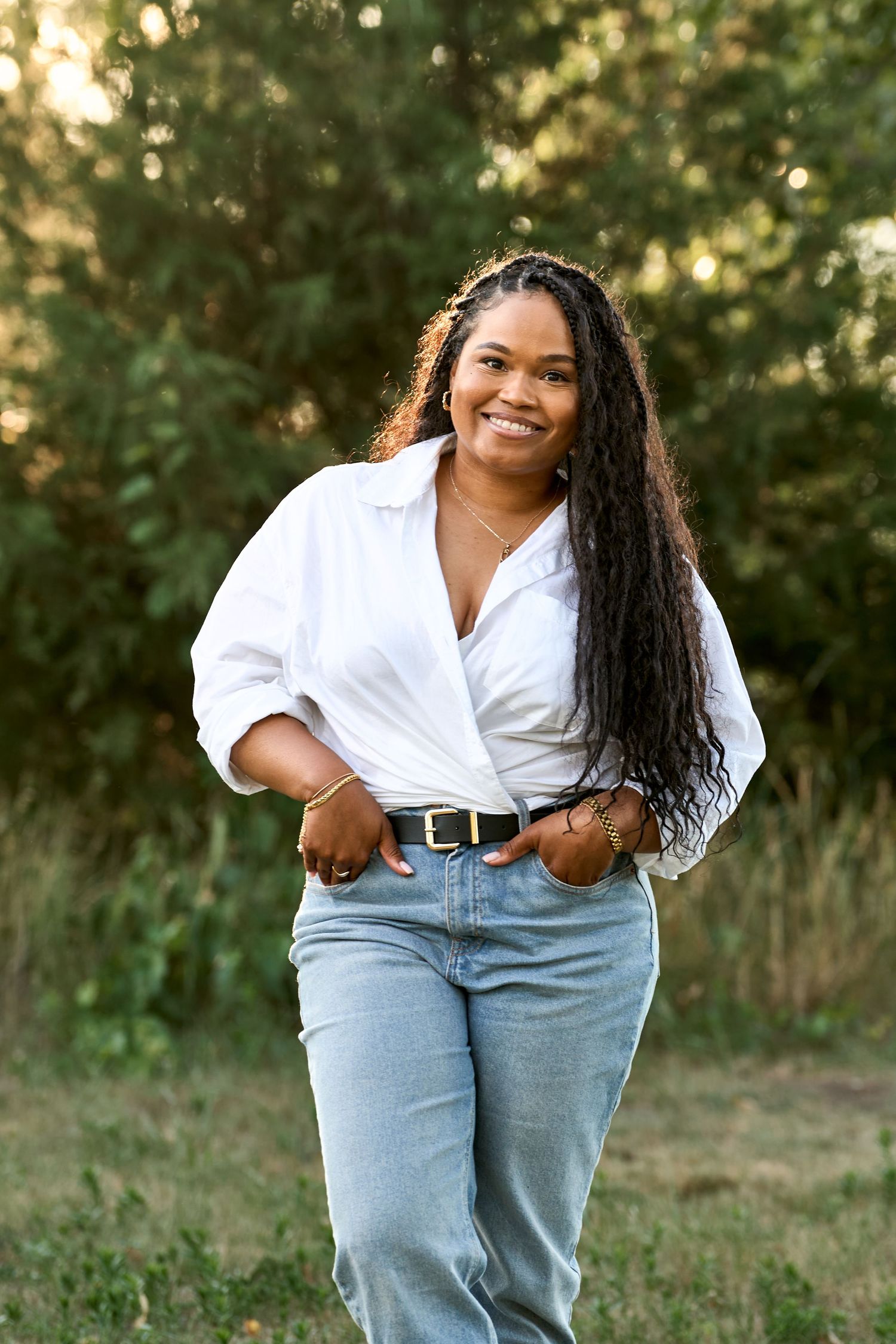 A person in a white button-down shirt and jeans poses outdoors with hands in pockets during golden hour.