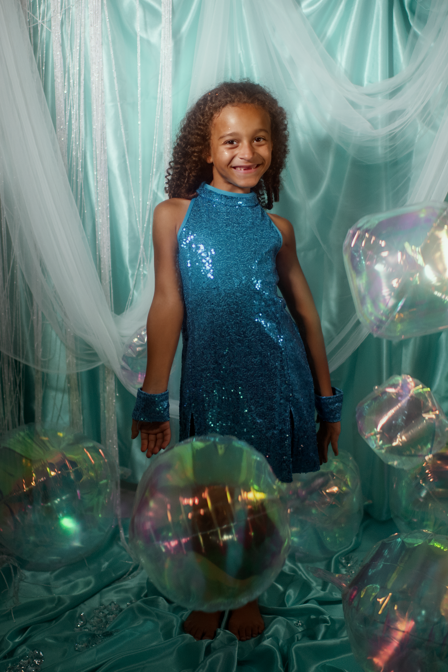 A smiling young girl in a teal sequin dress poses with iridescent bubbles against a mint green draped backdrop.