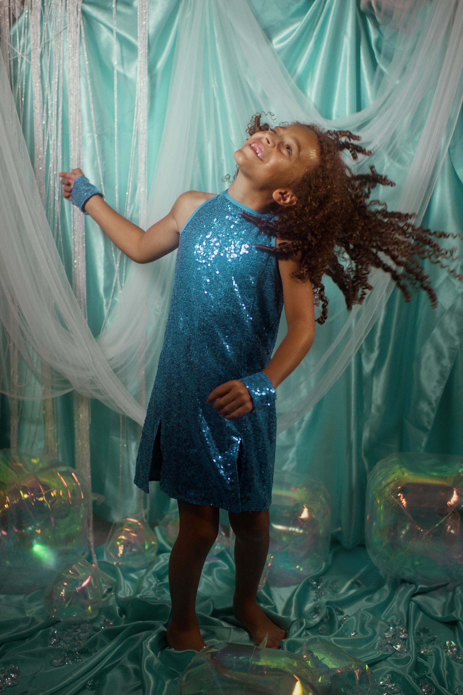 A young girl in a teal sequin dress dances joyfully with hair flying, surrounded by iridescent balloons and sheer curtains.