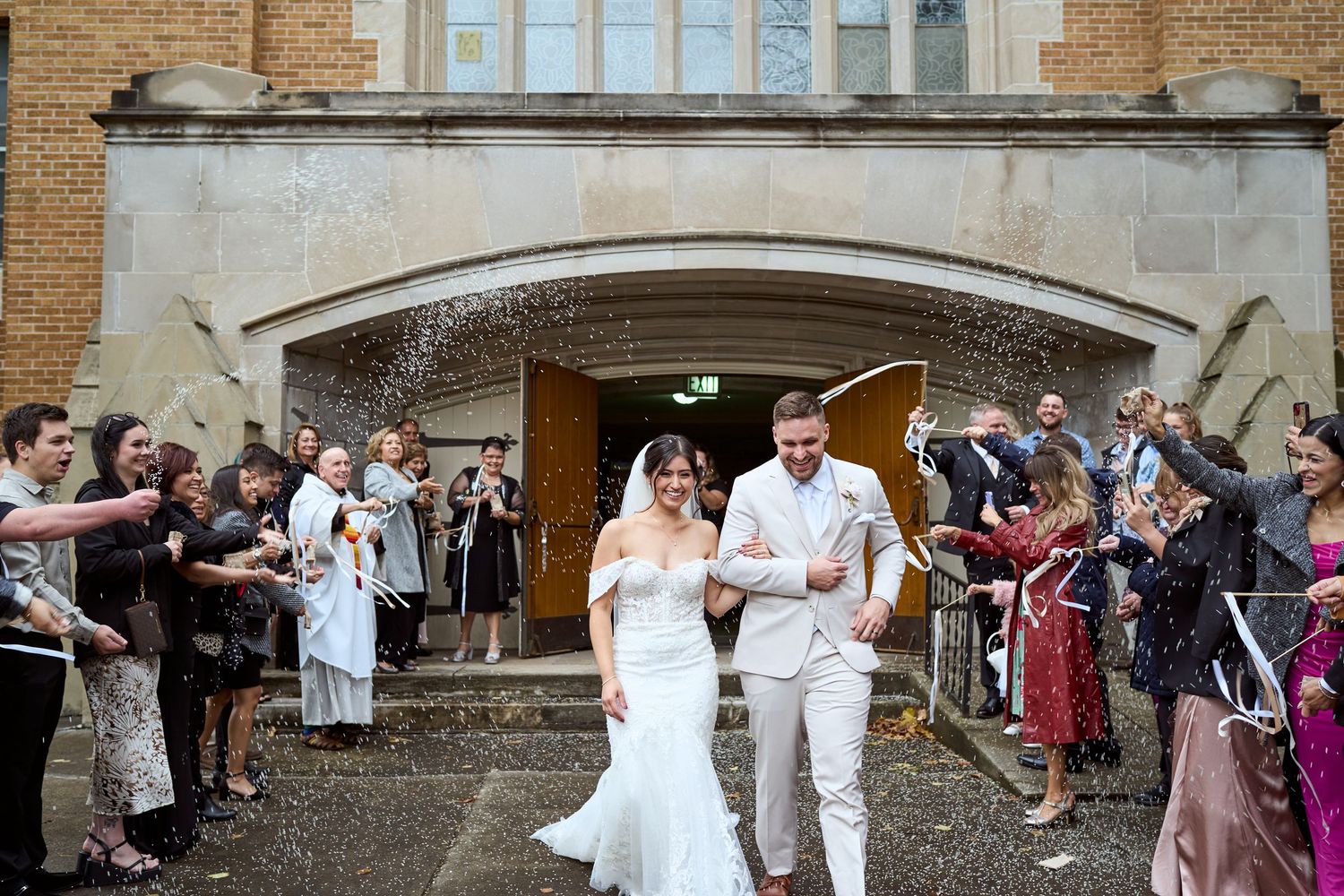 Wedding guests throw confetti at a couple during an outdoor celebration at a historic brick venue.