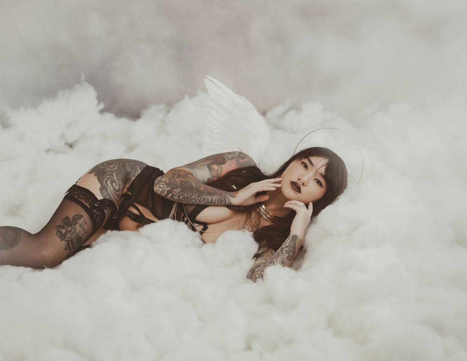 Ethereal figure lying in soft clouds wearing dark lingerie and tattoo sleeves in dreamy atmospheric photoshoot.