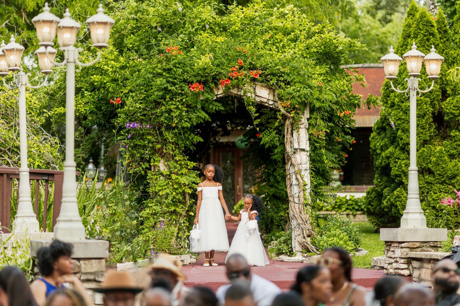 Outdoor wedding ceremony setup with white lamp posts and lush garden backdrop.