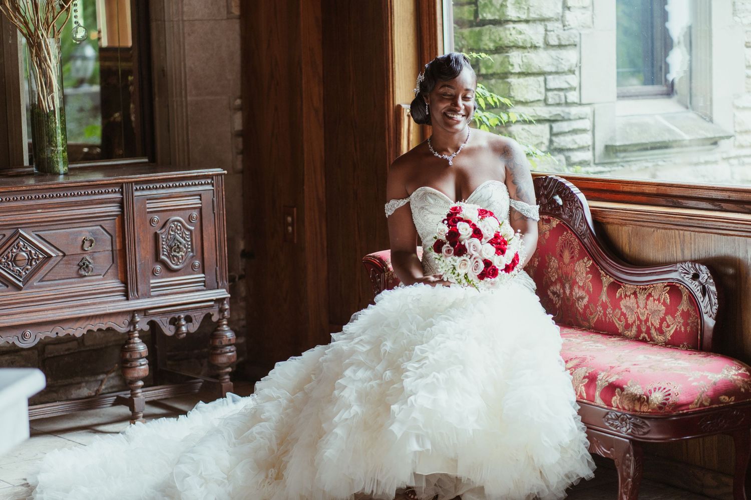Bride in off-shoulder ballgown poses on vintage red sofa holding red and white bouquet.