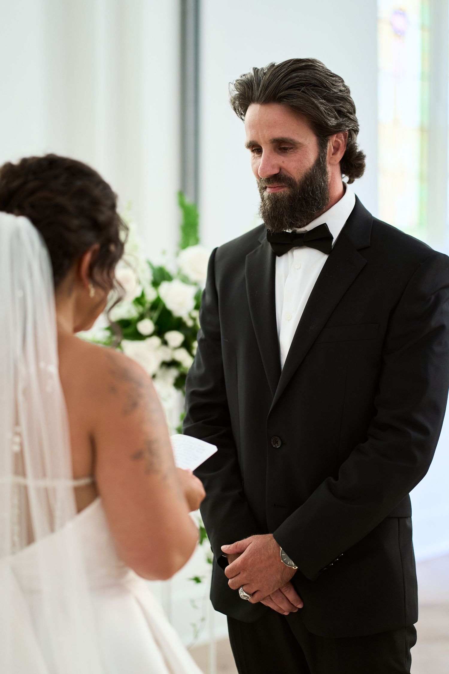 A couple exchanges vows during an intimate wedding ceremony with white floral decorations in the background.