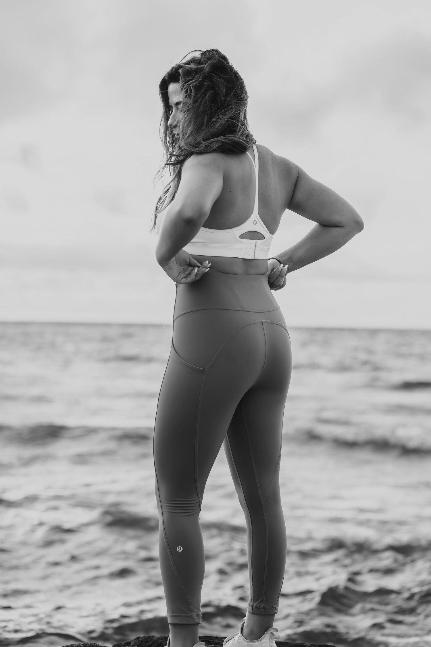 Black and white photo of woman in athletic wear standing by ocean waves.