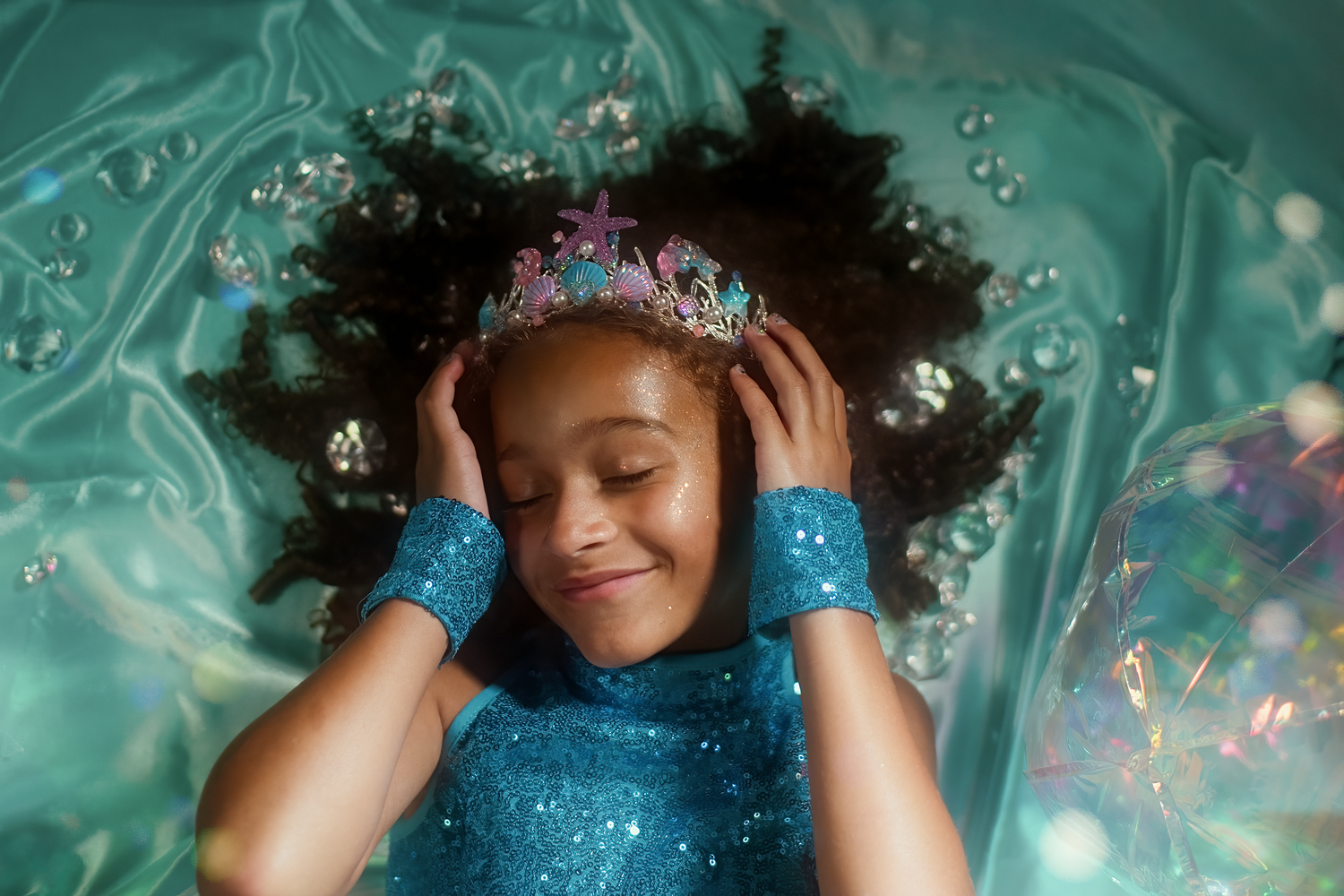 A young Black girl wearing a blue sequin dress and crown lies on teal fabric surrounded by bubbles, smiling with eyes closed.