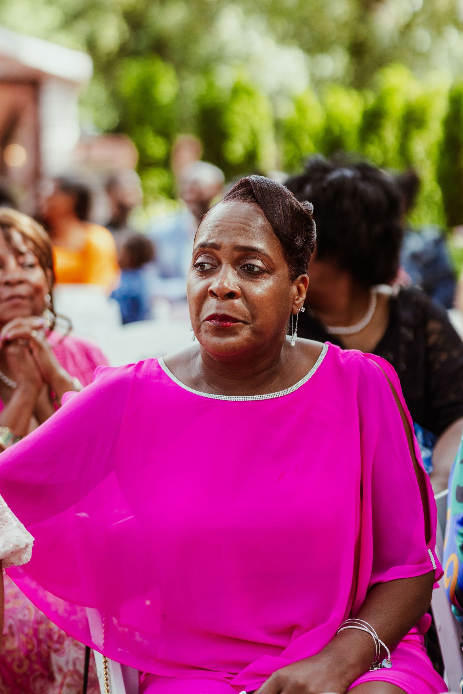 Wedding guest wearing vibrant pink flowing top watches ceremony in outdoor garden setting.