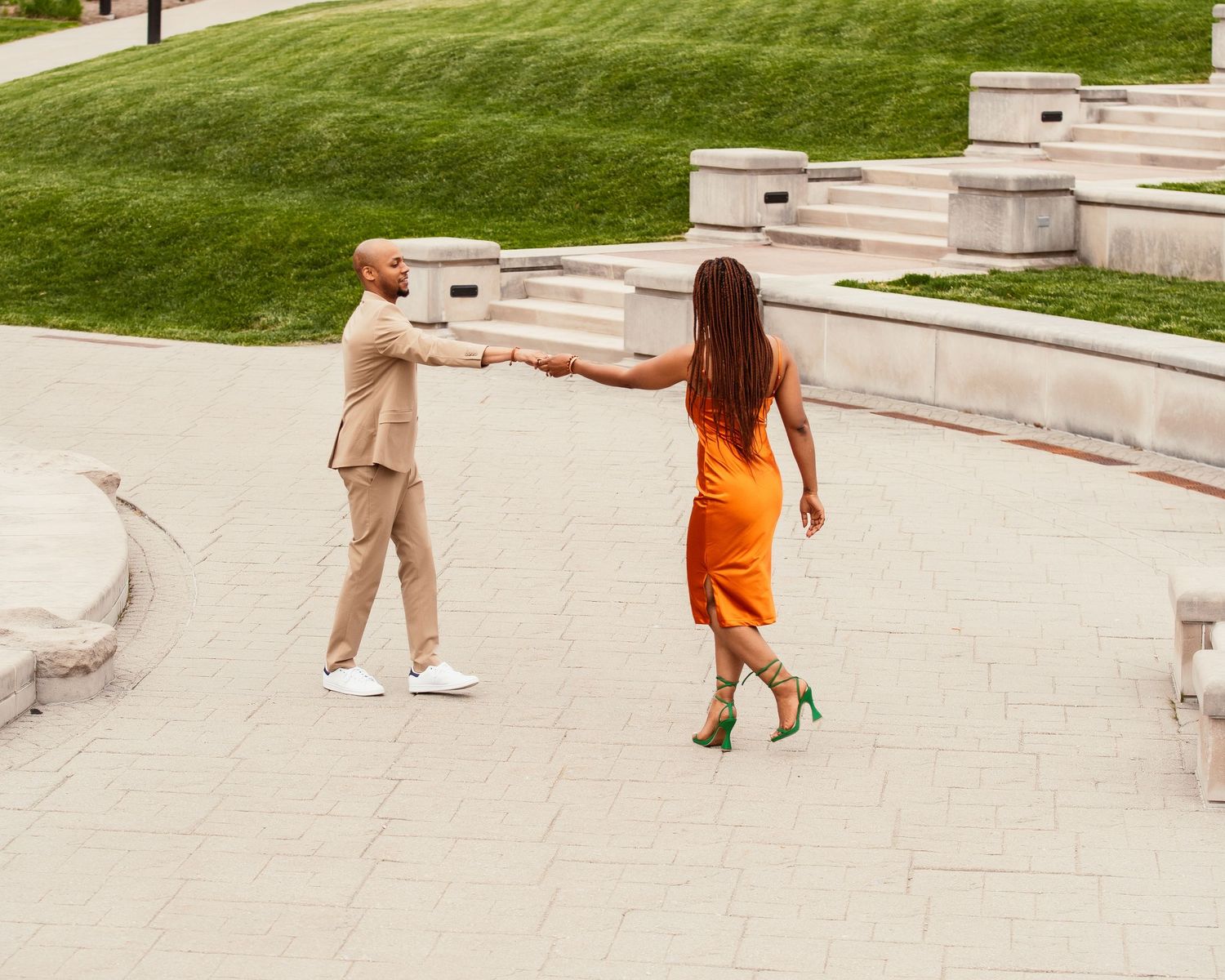 Couple in orange dress and beige suit dancing on concrete steps with manicured lawn in background.