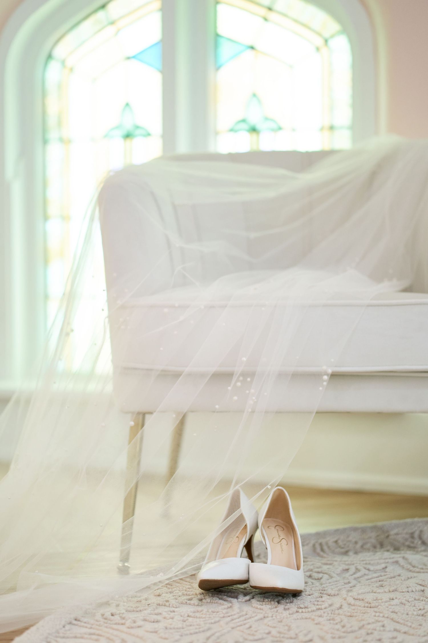 Elegant white bridal shoes sit on a patterned rug near a window with stained glass details.