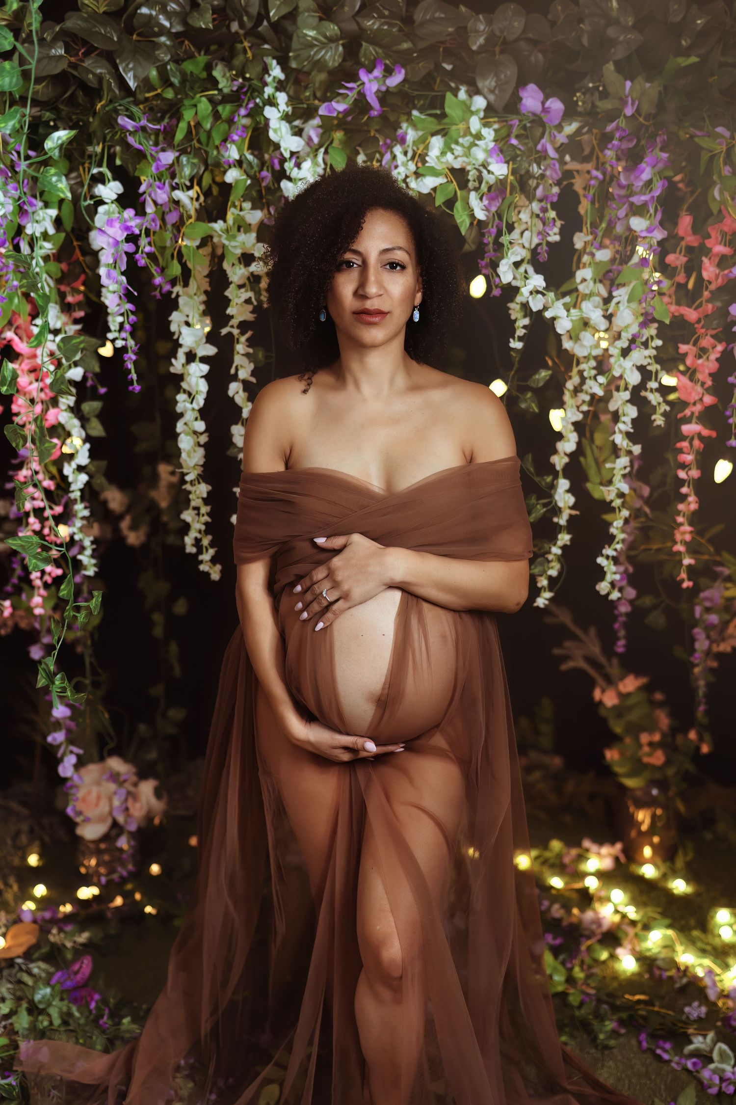 Maternity photoshoot in brown sheer gown against enchanted garden backdrop with hanging floral arrangements and soft lighting.