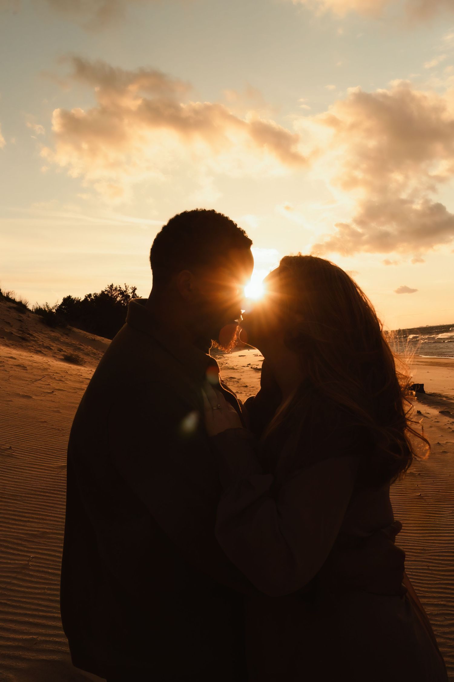 A silhouetted couple shares an intimate moment at sunset on sand dunes with golden clouds in the background.