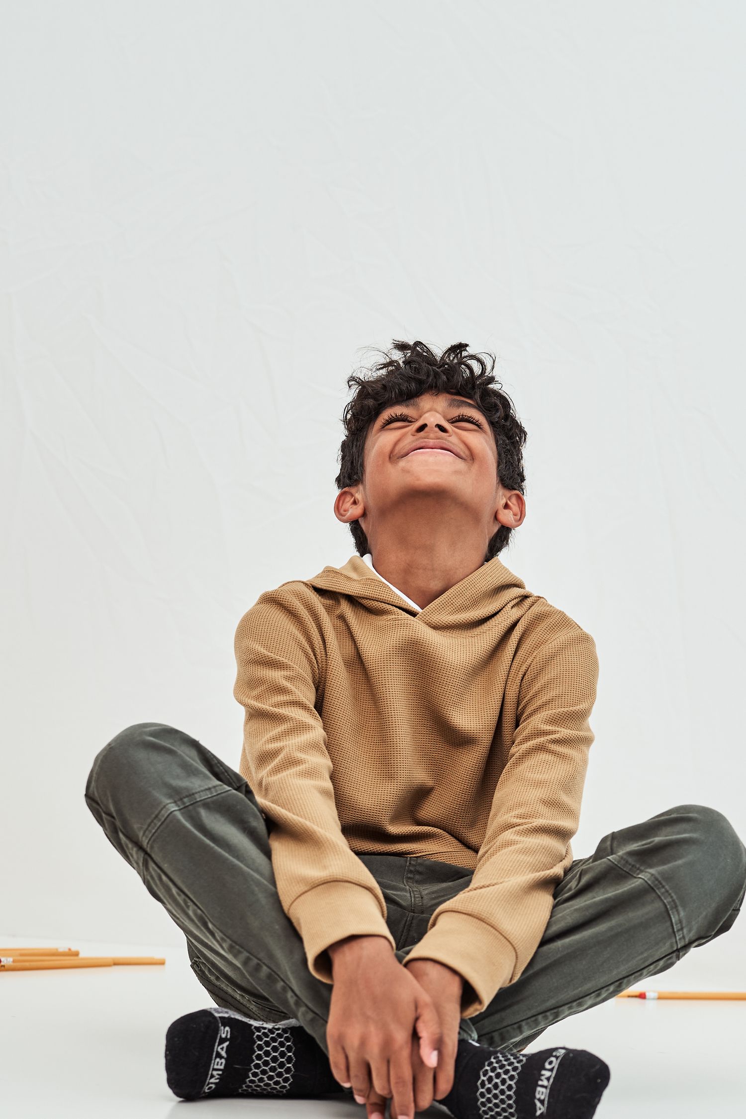 A young person in a tan hoodie and cargo pants sits cross-legged while looking up and smiling against a white background.