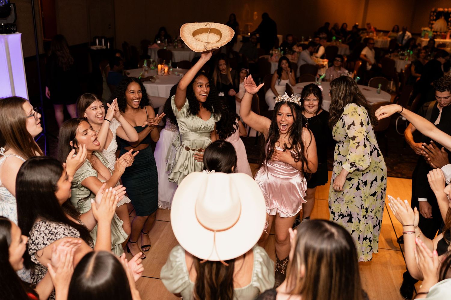 Energetic crowd wearing dresses and cowboy hats dances together on a dark dance floor at a party celebration.