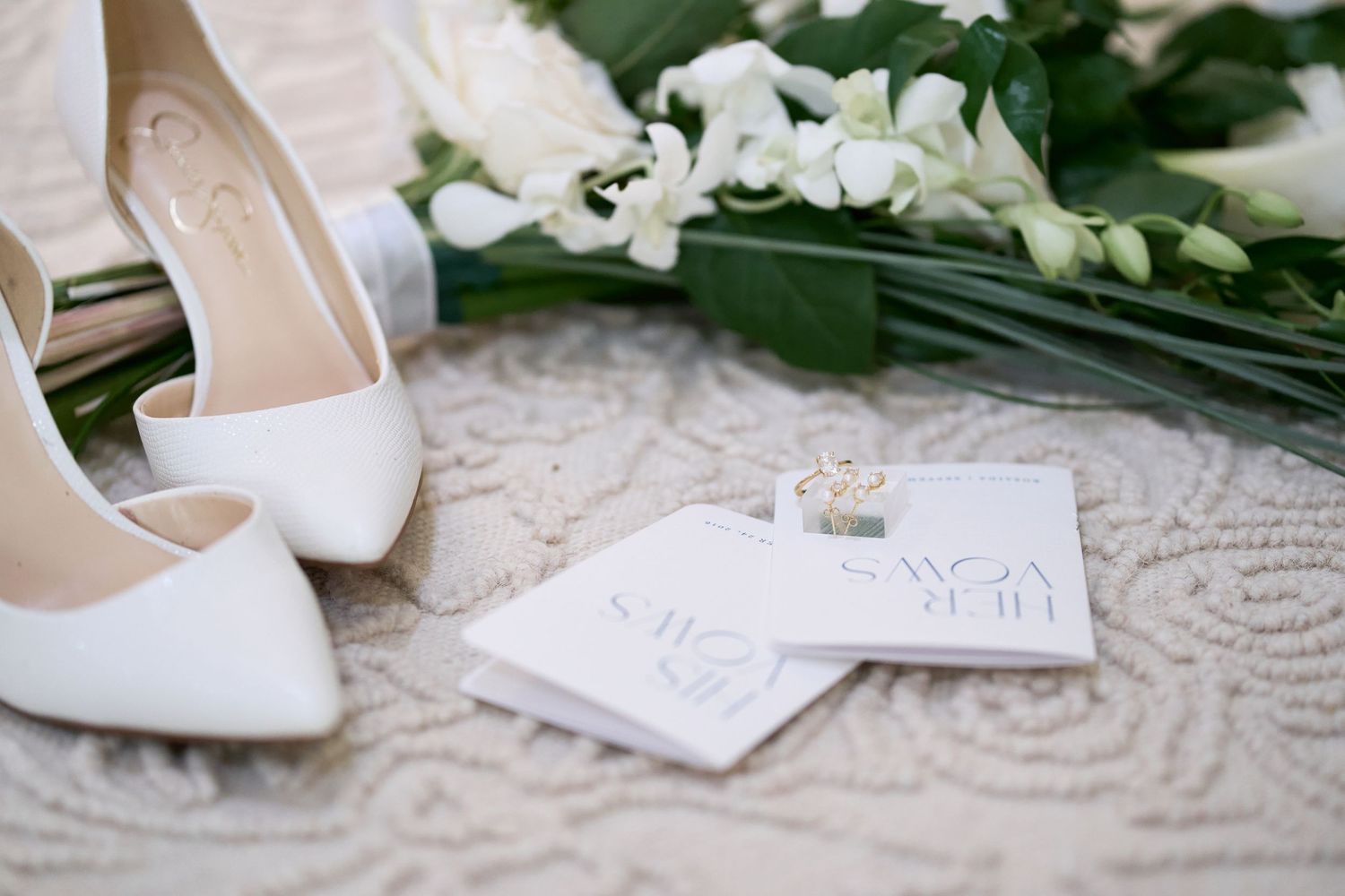White wedding shoes placed next to handwritten notes and white floral arrangements on a lace surface.