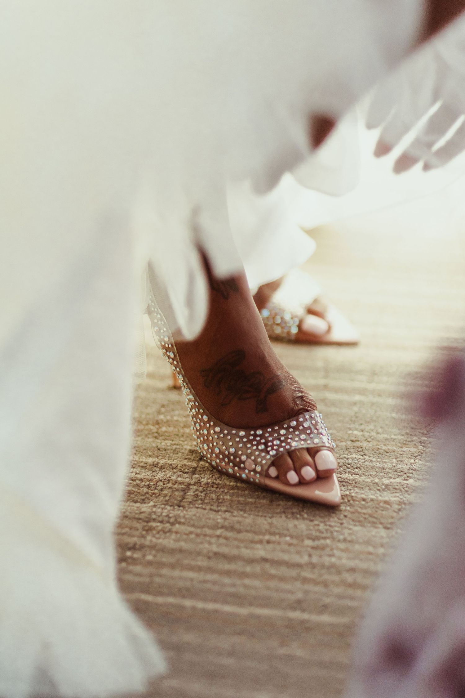 Detailed shot of crystal-embellished bridal shoes with white tulle dress flowing around them.