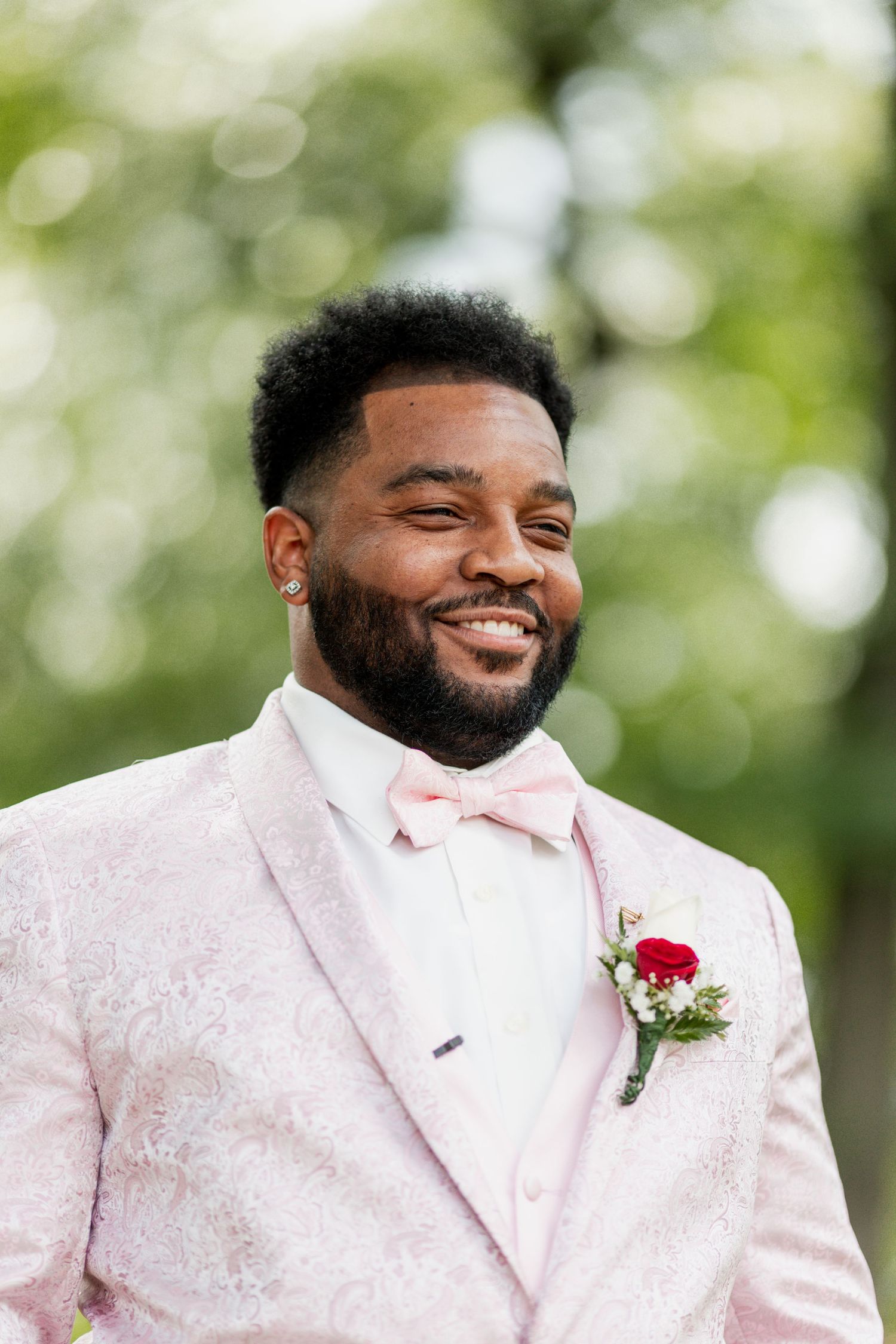 Someone wearing a pink tuxedo jacket and bow tie smiles brightly with a red boutonniere.