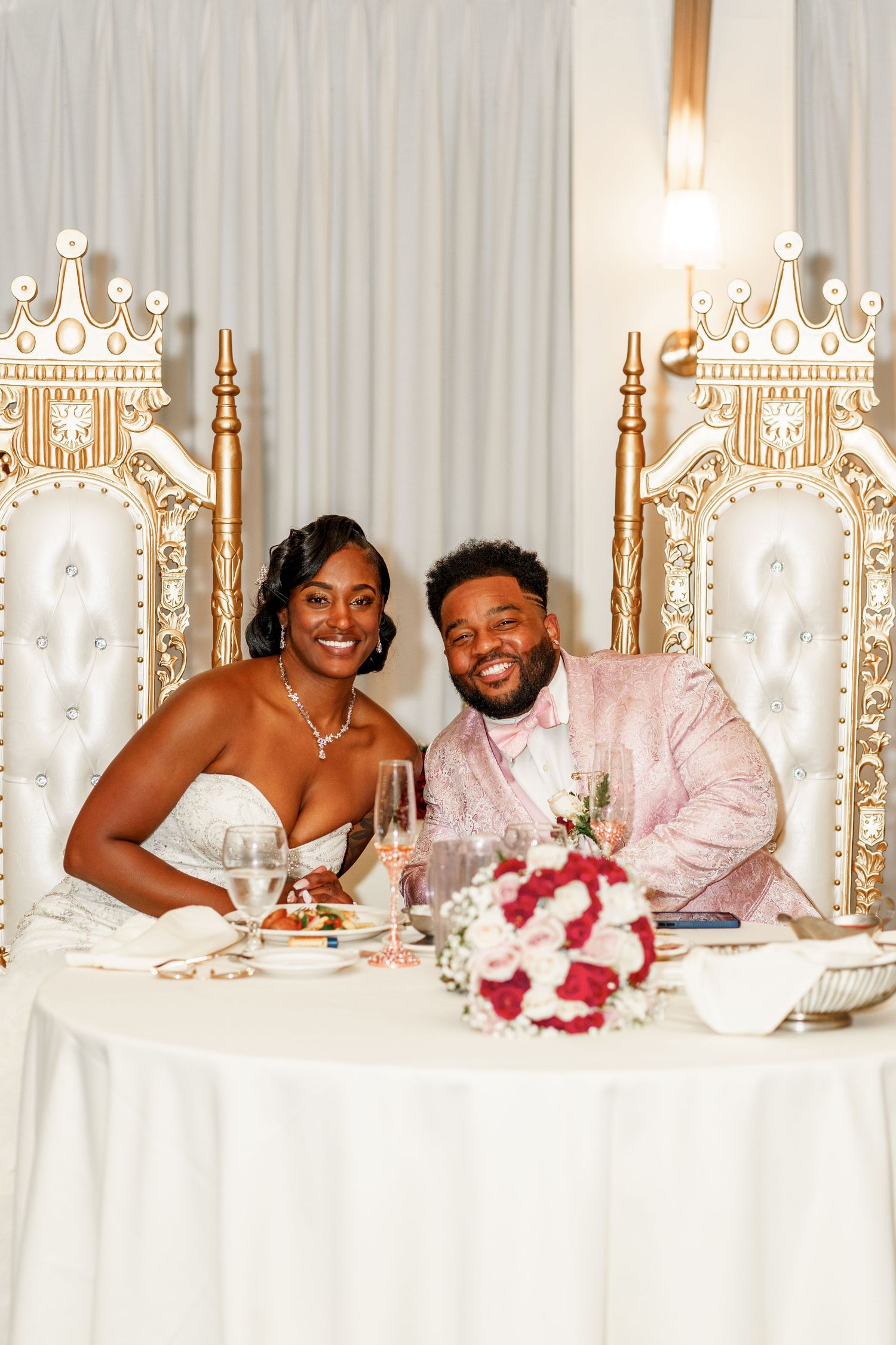 Newlyweds share intimate moment at reception sweetheart table with ornate throne chairs.