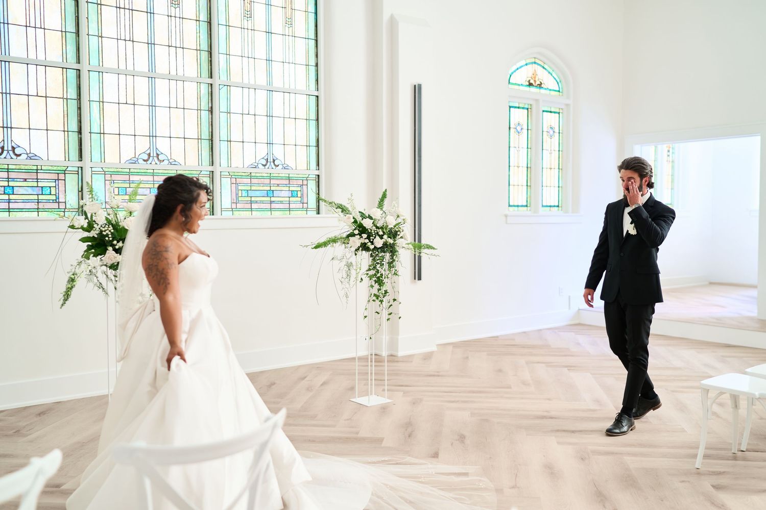 A bride in a white gown stands in a bright chapel-like room with stained glass windows while a photographer captures the moment.