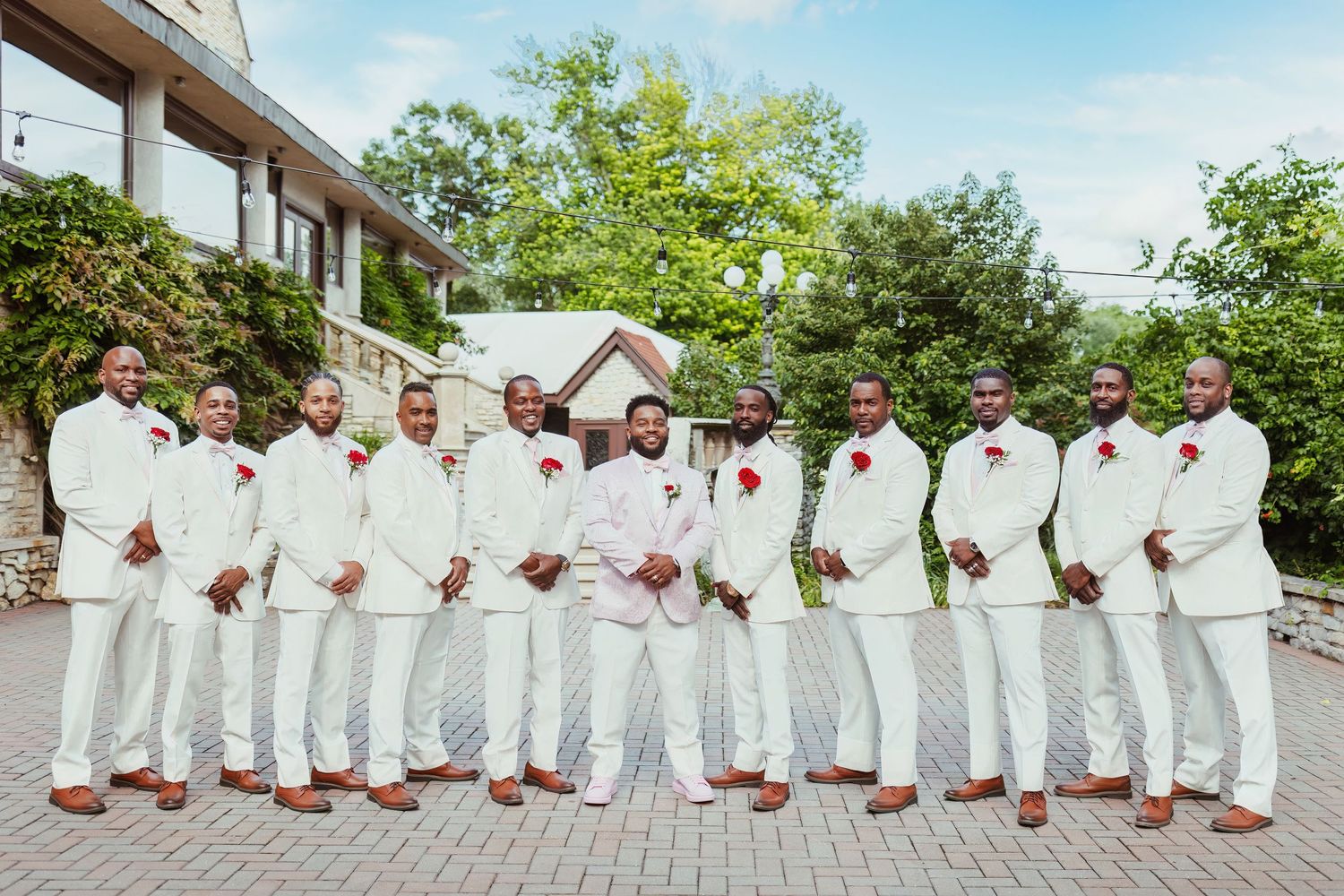 Wedding party in all-white suits with brown shoes pose outdoors on stone patio.