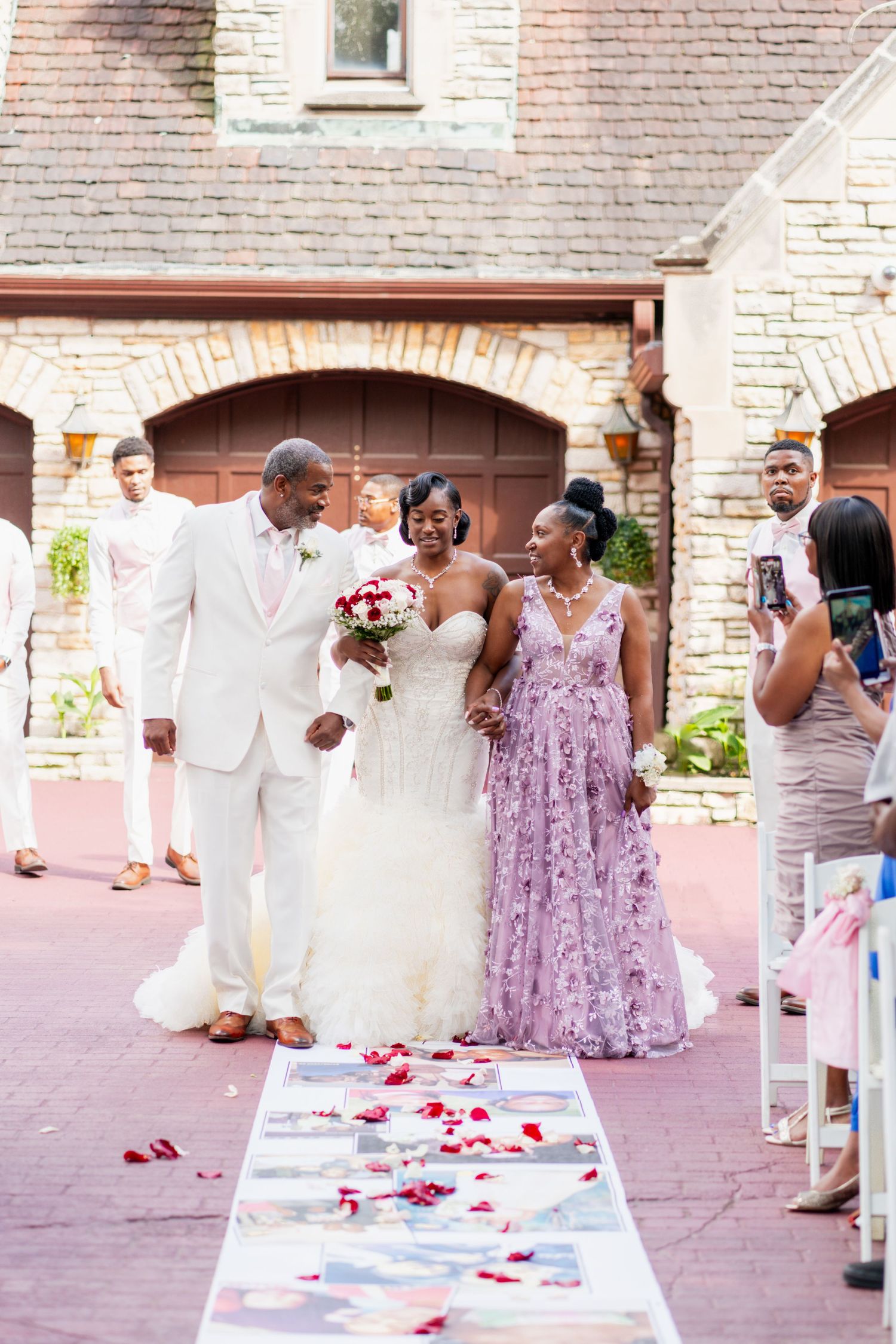 Wedding party members walk down a petal-strewn aisle lined with guest chairs.