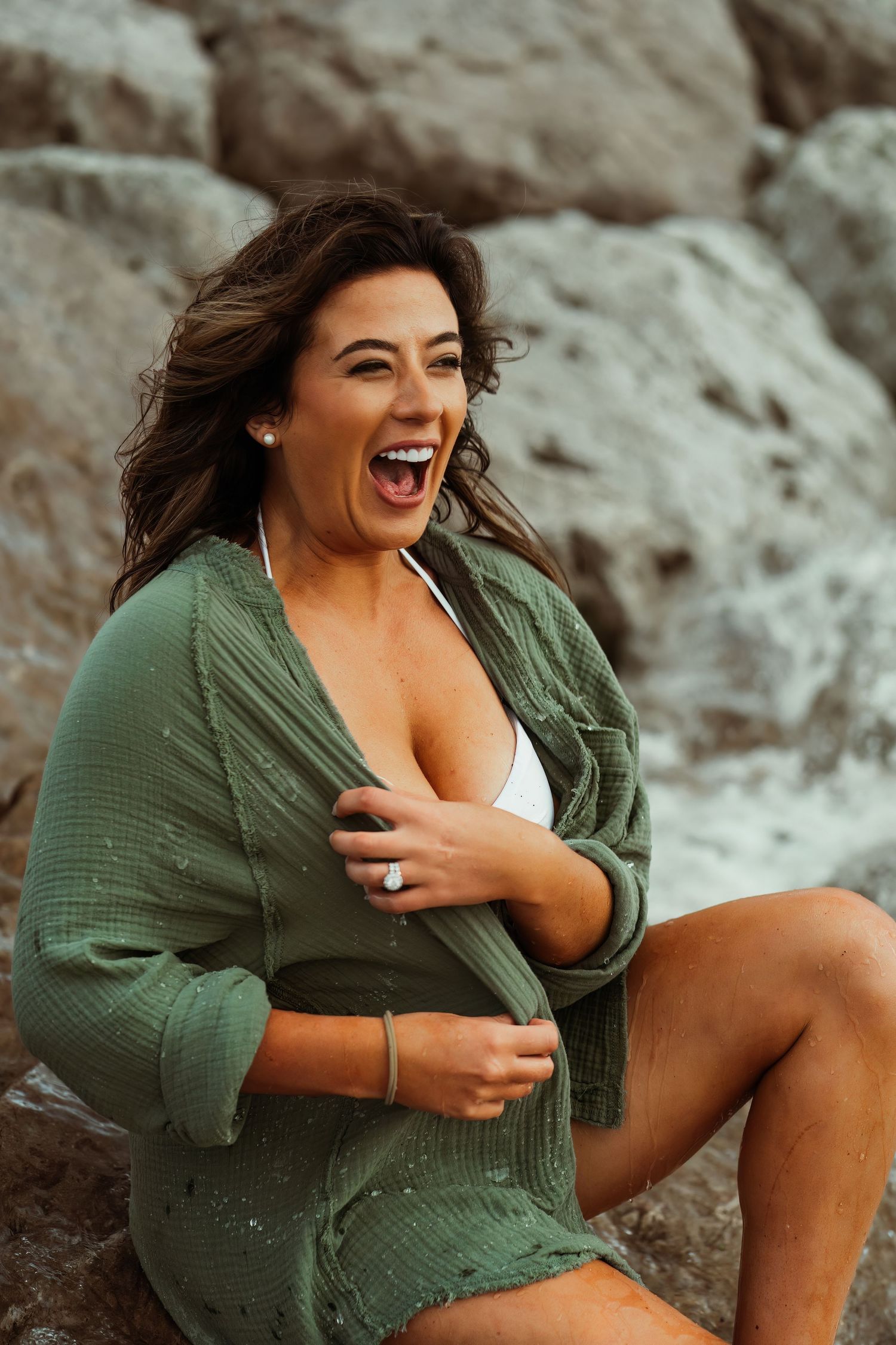 Woman wearing green cardigan laughs joyfully while posing on rocky beachside.