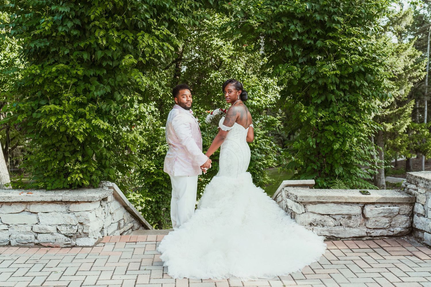 Newlywed couple holds hands on stone pathway surrounded by green foliage.